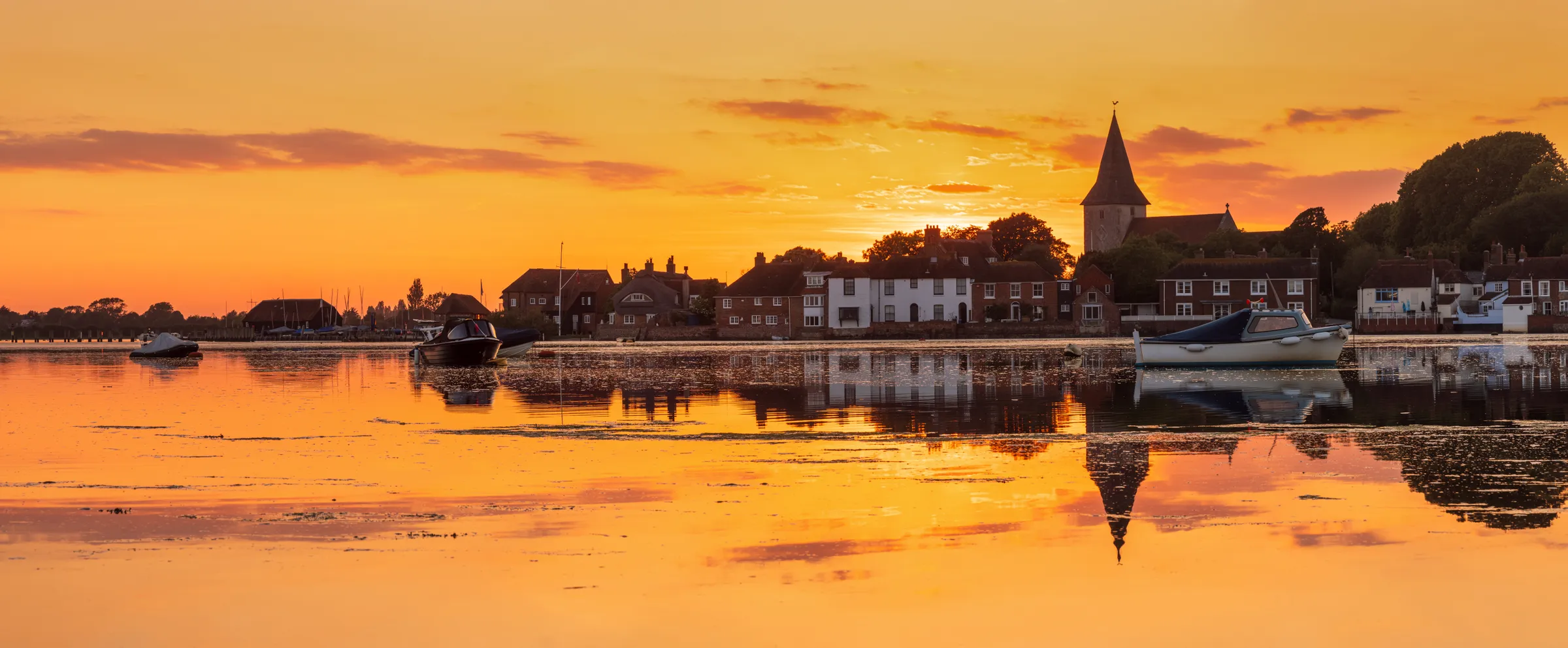 A vivid orange sunset over Bosham Quay with moored boats, waterfront houses and Bosham Church reflected in still harbour water.