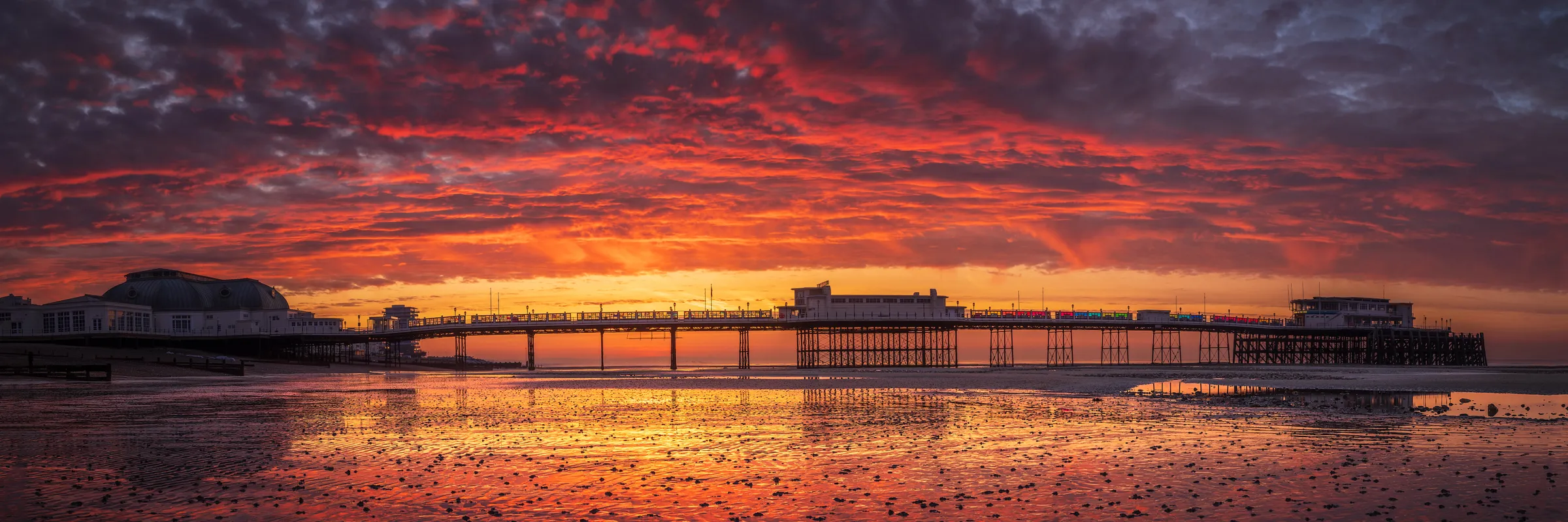 A dramatic red and orange sunrise stretching across the sky behind Worthing Pier, reflected in wet sand and low tide textures on the Sussex coast.