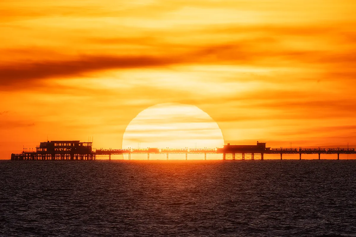 The setting sun perfectly aligned behind Worthing Pier, appearing oversized due to compression, with the structure silhouetted against a deep orange sky.