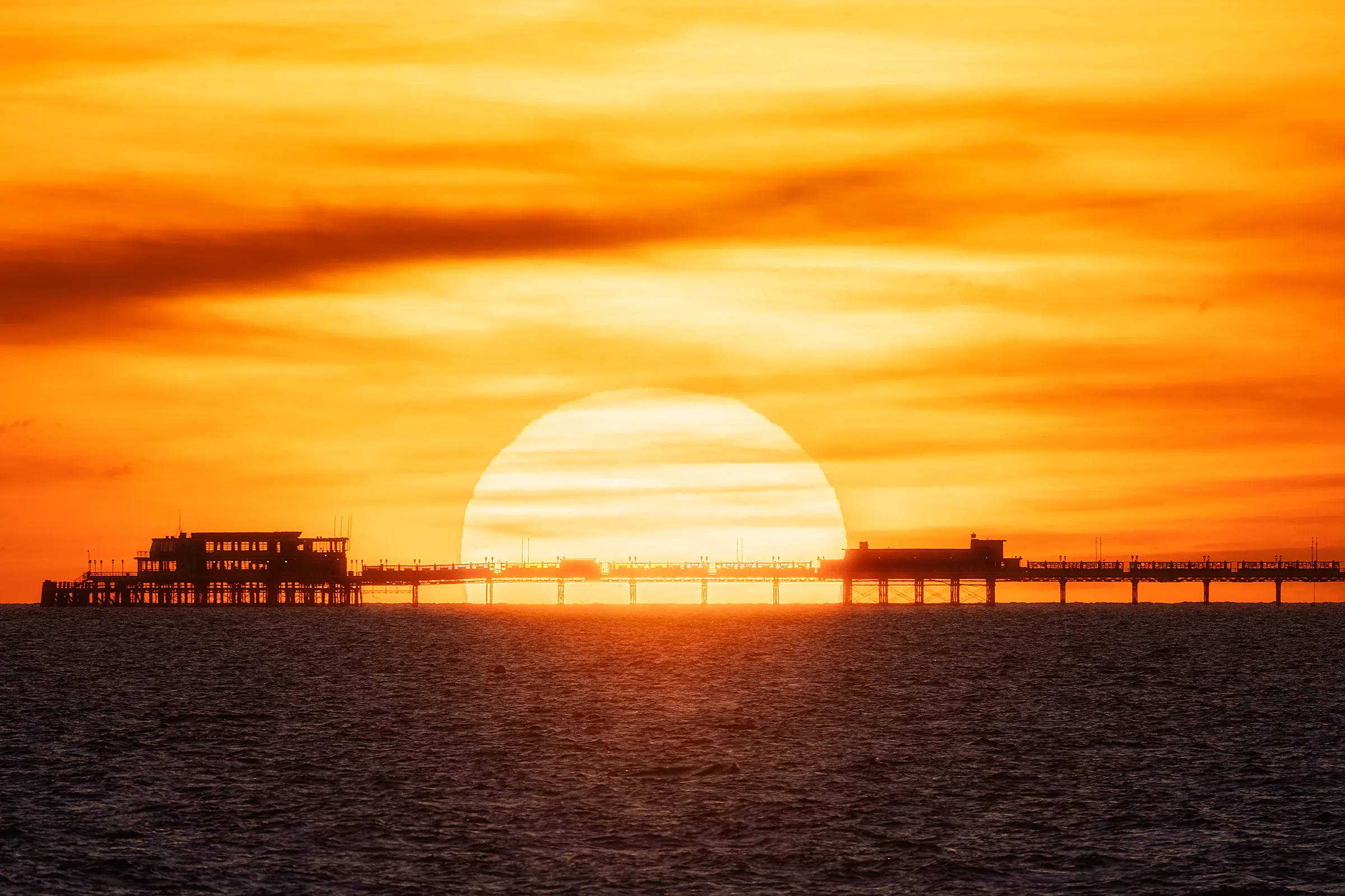 The setting sun perfectly aligned behind Worthing Pier, appearing oversized due to compression, with the structure silhouetted against a deep orange sky.