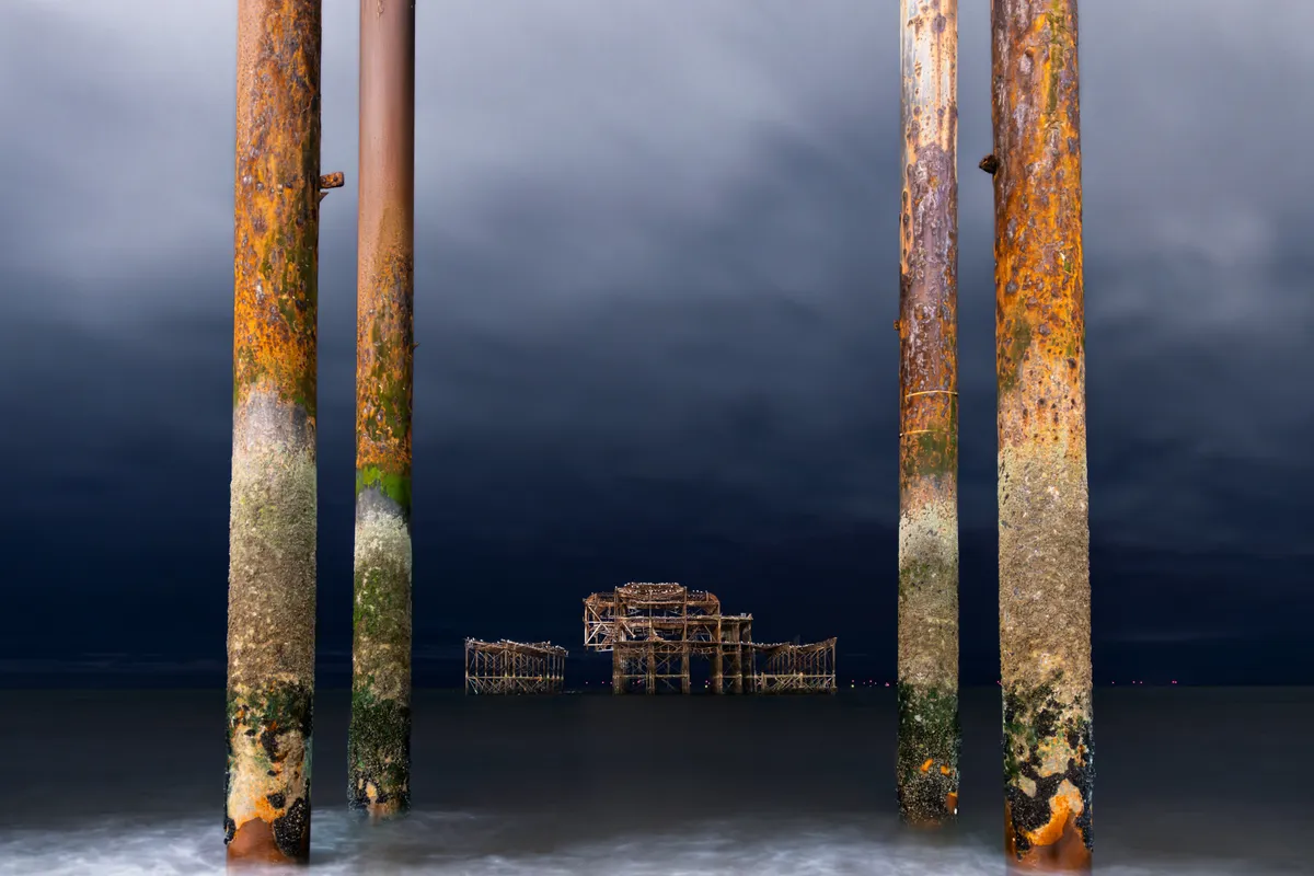The remains of Brighton’s West Pier framed between rusted foreground pillars, with textured decay contrasting against a dark coastal sky.