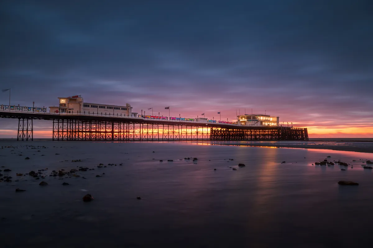 Worthing Pier at dawn with soft pastel light, illuminated pier structures, and a long exposure smoothing the water with scattered foreground detail.
