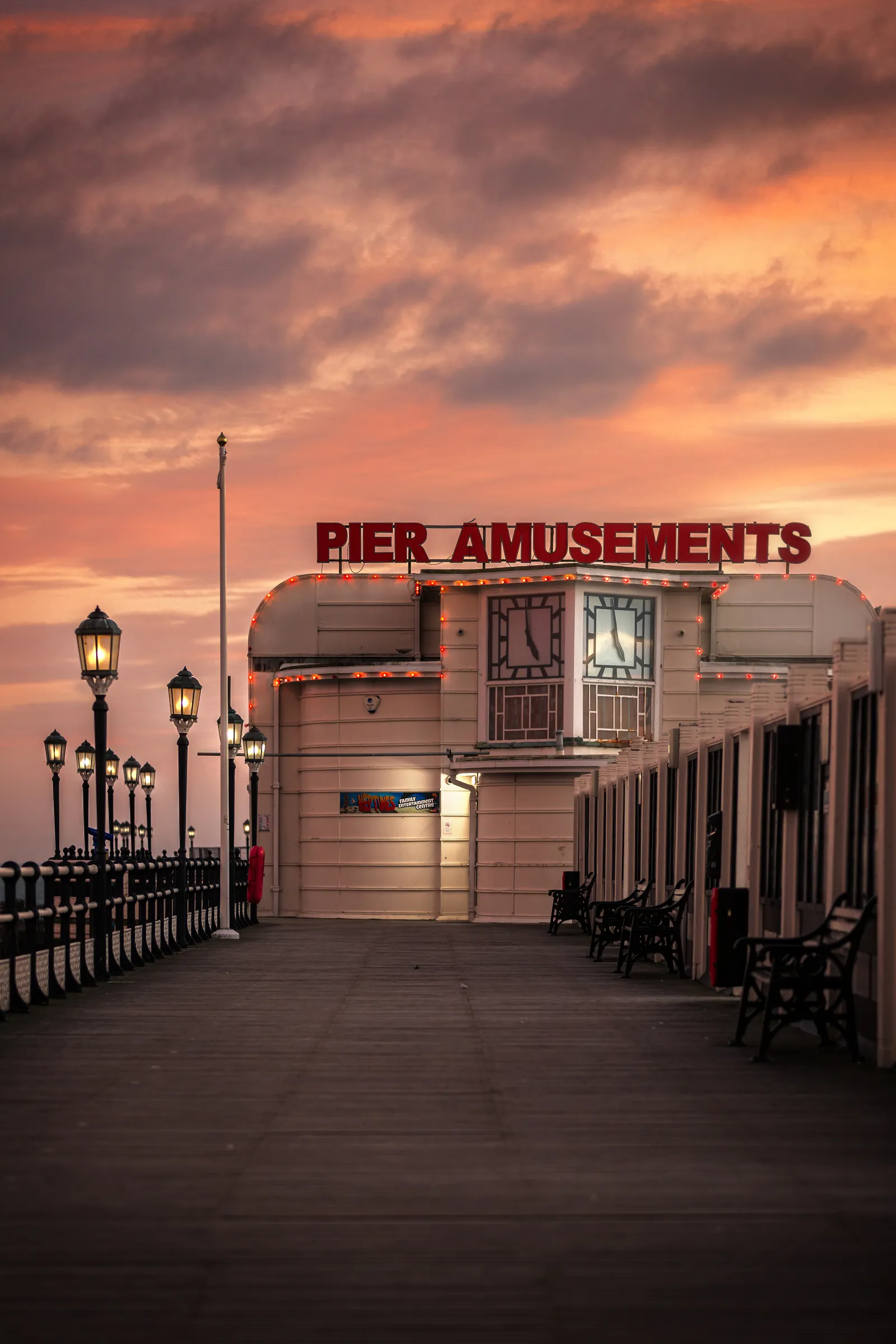 Worthing Pier amusements building at sunset, framed by lampposts along the boardwalk, with warm pastel skies and soft evening light.