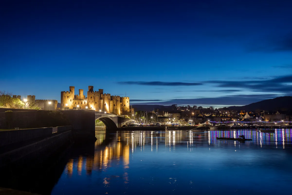 Conwy Castle illuminated at blue hour beside the harbour, with warm lights reflecting across calm water under a deep blue evening sky.