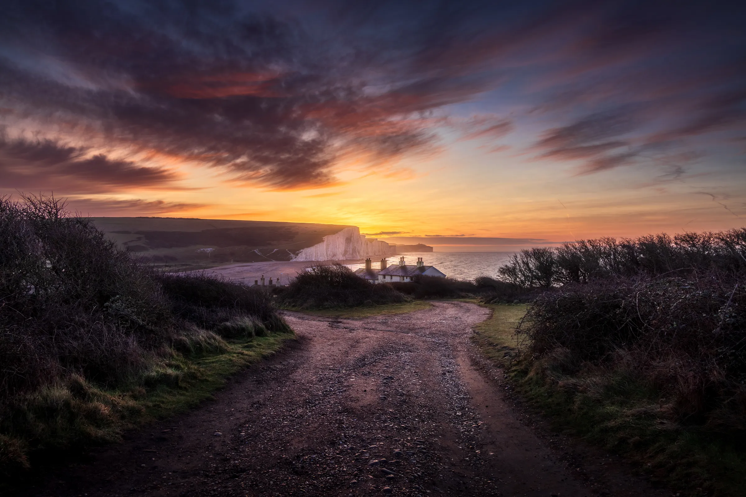 A winding track leading to the Coastguard Cottages at Cuckmere Haven at sunrise, with golden light over the Seven Sisters cliffs and soft cloud detail.