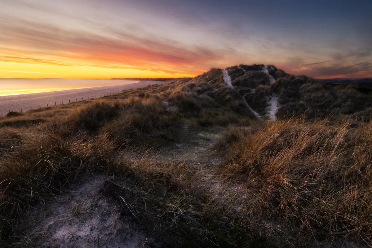 Coastal sand dunes at Littlehampton at sunset, with winding footpaths through grasses leading toward a glowing horizon over the sea.