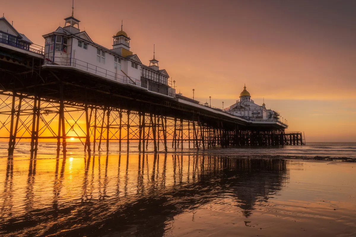 Eastbourne Pier at sunrise with the sun glowing beneath the structure, casting warm reflections across wet sand and the ironwork silhouetted above.