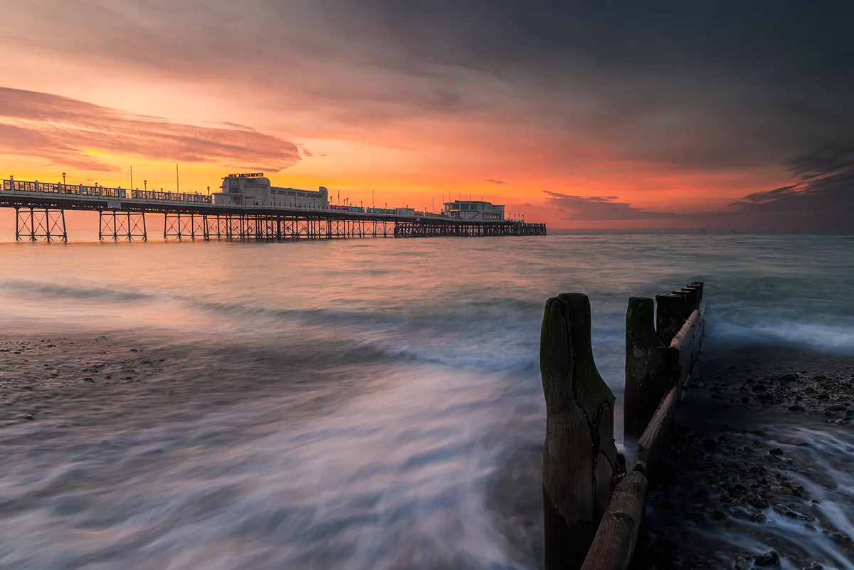 Worthing Pier at sunset with waves sweeping around a wooden groyne, warm orange sky reflecting across moving water.