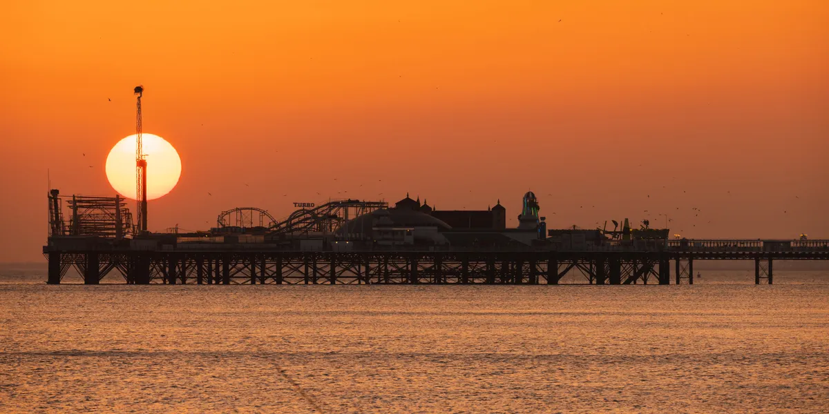 The sun setting behind Brighton Palace Pier, aligned with a tower and silhouetting the amusement rides against a vivid orange sky.