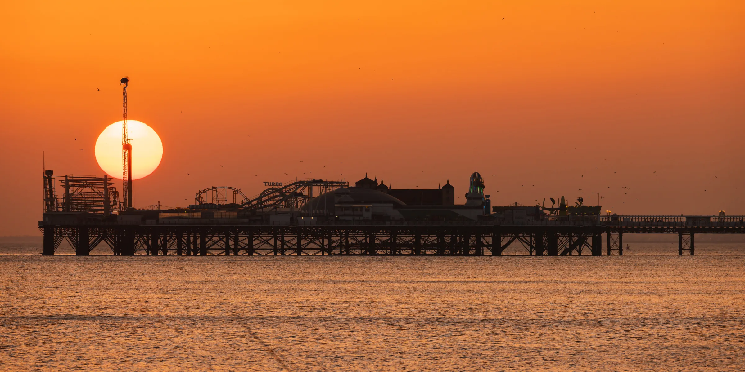 The sun setting behind Brighton Palace Pier, aligned with a tower and silhouetting the amusement rides against a vivid orange sky.
