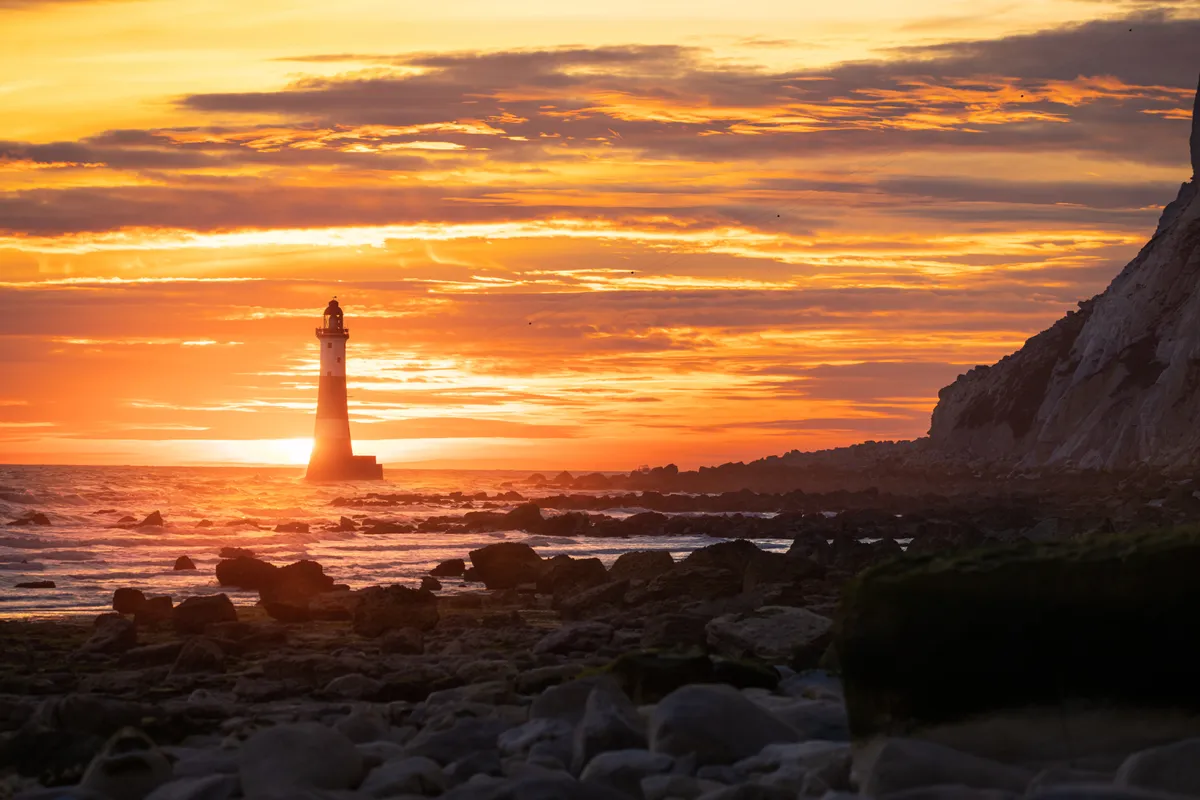 Beachy Head Lighthouse silhouetted against a vivid orange sunrise, with waves breaking over rocks and cliffs framing the coastline.
