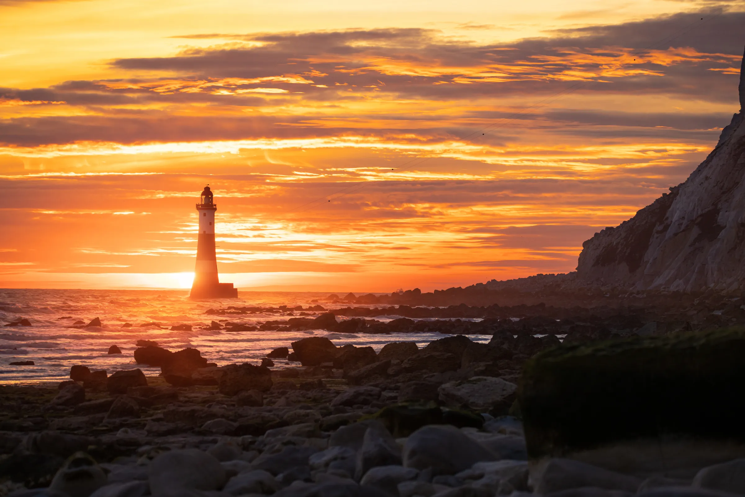 Beachy Head Lighthouse silhouetted against a vivid orange sunrise, with waves breaking over rocks and cliffs framing the coastline.