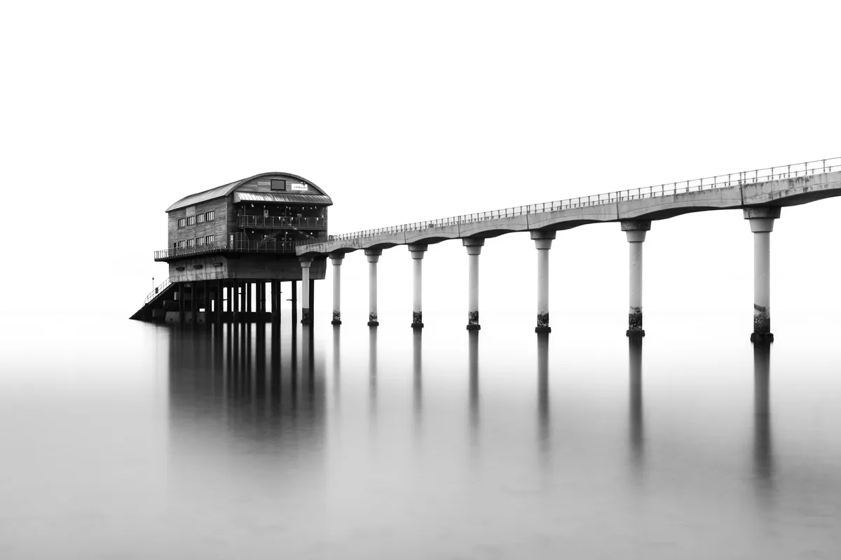 Bembridge Pier in minimalist black and white, isolated against smooth long-exposure water with repeating columns fading into stillness.
