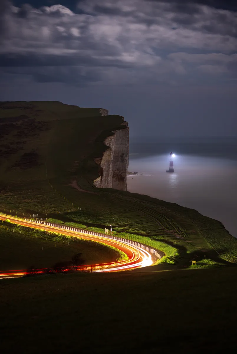 Beachy Head cliffs at night with a winding road captured as light trails, leading toward the lighthouse glowing through mist over the sea.