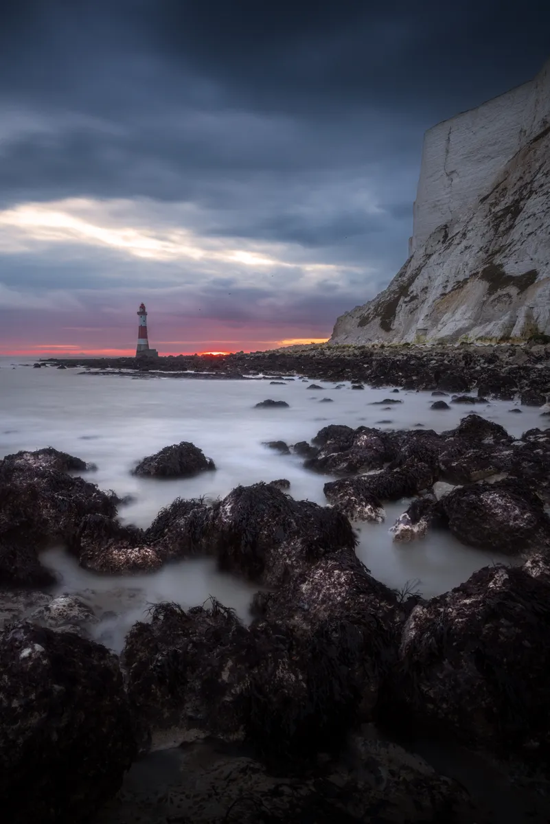 Beachy Head cliffs and lighthouse at dusk, with soft long-exposure water and dark foreground rocks under a heavy sky.