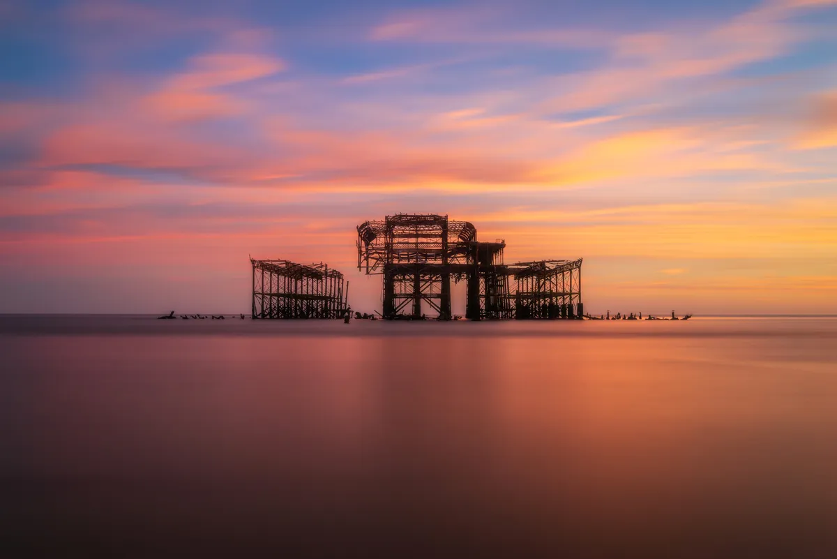 The skeletal remains of Brighton West Pier silhouetted against a pastel sunset, with smooth long-exposure water creating a soft, minimal foreground.
