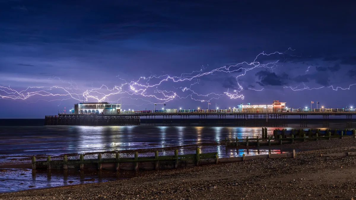 Lightning forks across the sky above Worthing Pier at night, reflecting across wet sand and the calm sea below.