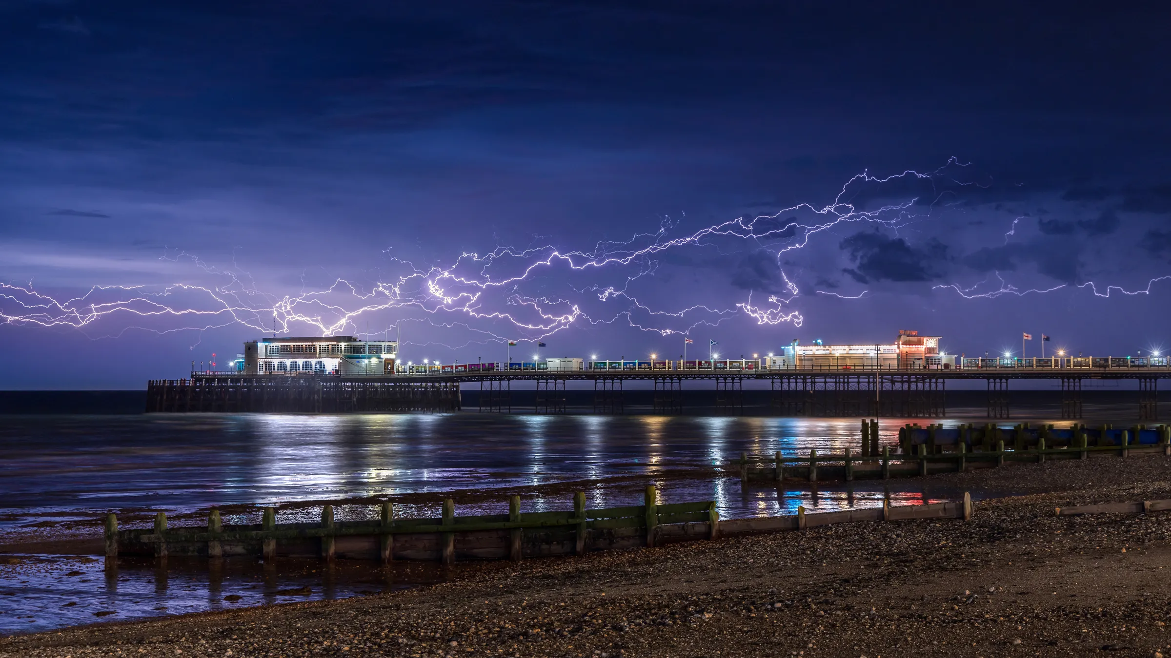 Lightning forks across the sky above Worthing Pier at night, reflecting across wet sand and the calm sea below.
