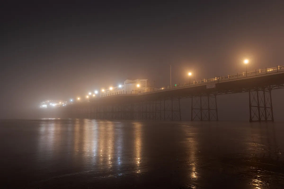 Worthing Pier disappearing into dense fog at night, with warm lights reflecting across wet sand and fading into the distance.