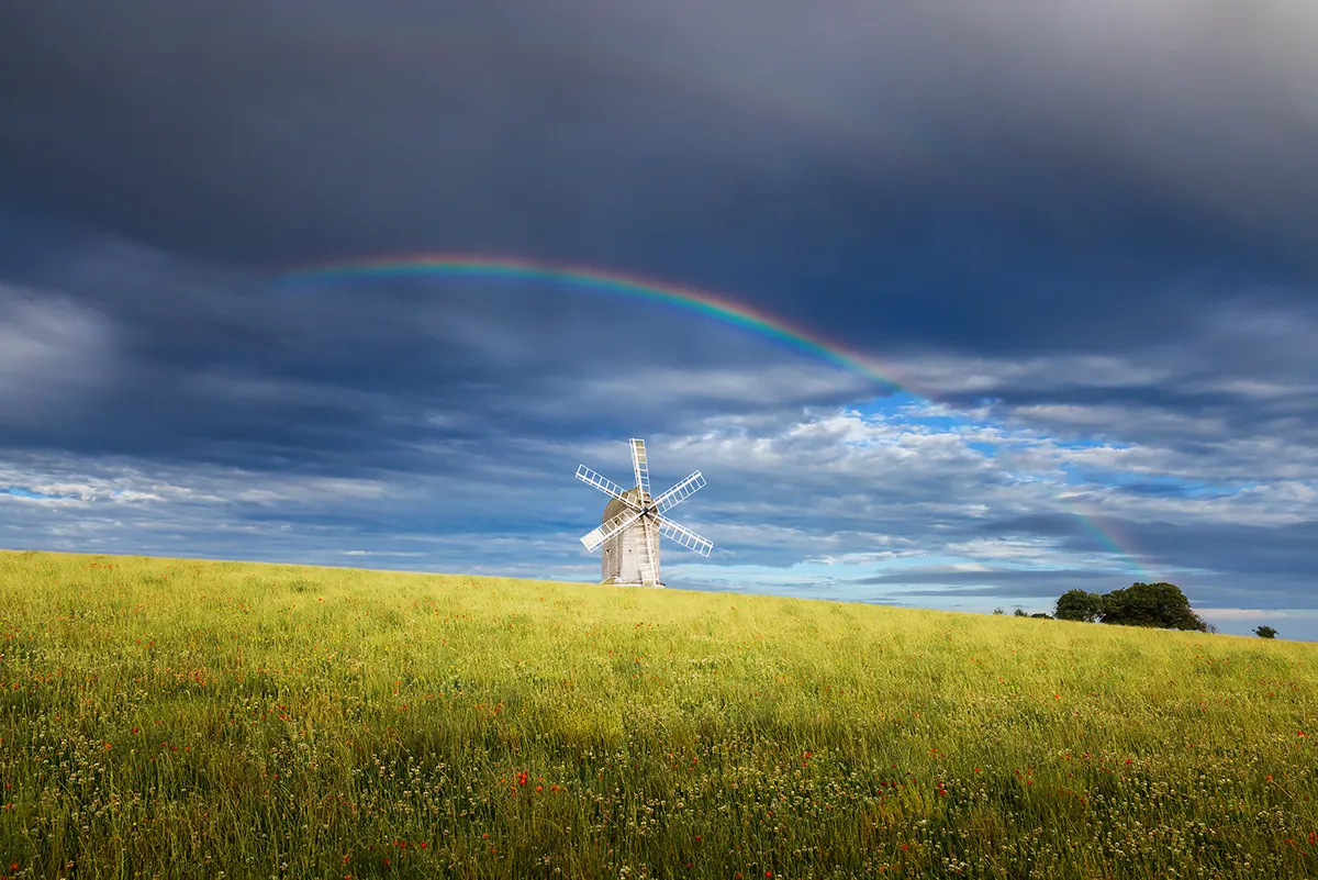 Ashcombe Windmill stands beneath a dramatic divided sky, with a rainbow crossing storm clouds above a sunlit summer field in Kingston, East Sussex.