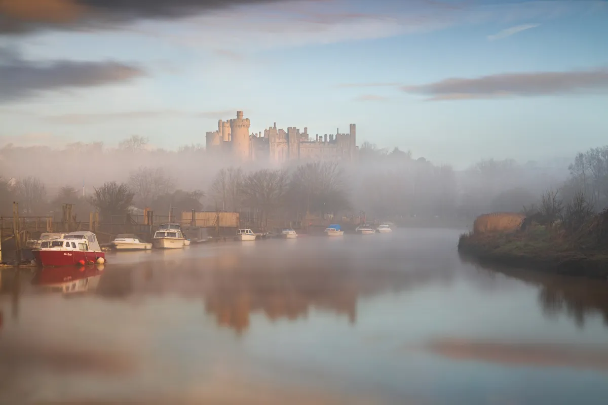 Arundel Castle rising above mist along the river with boats reflected in still water at sunrise.