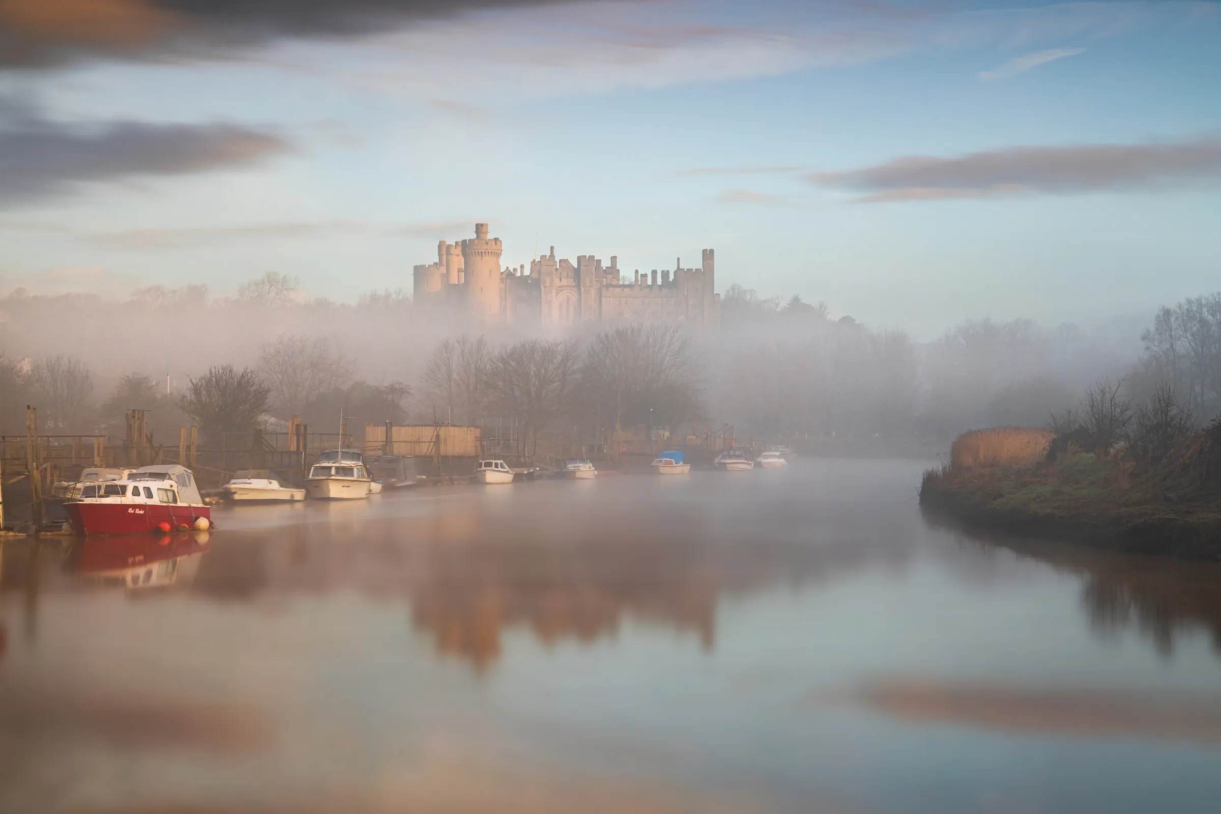 Arundel Castle rising above mist along the river with boats reflected in still water at sunrise.
