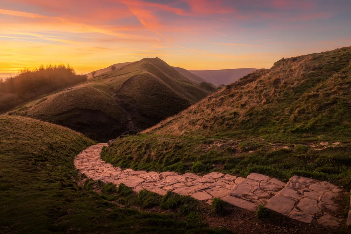 Rolling hills in the Peak District at sunset with a winding stone path leading through the landscape under warm orange skies.