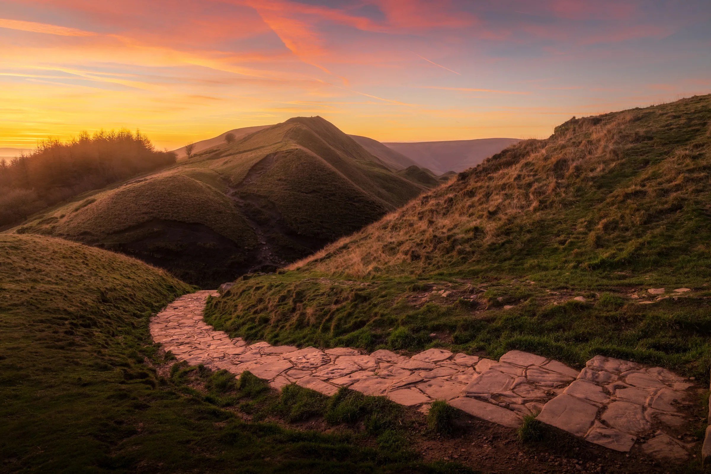 Rolling hills in the Peak District at sunset with a winding stone path leading through the landscape under warm orange skies.