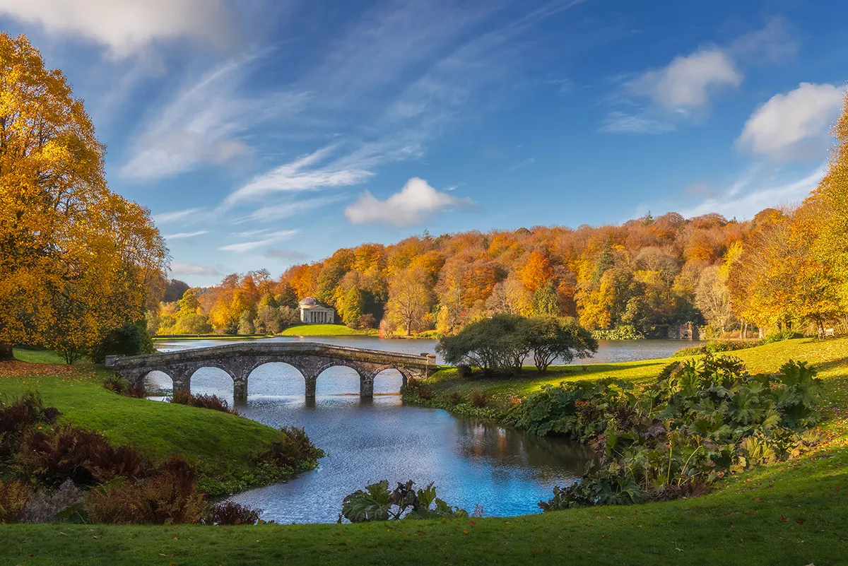 A stone bridge crossing a calm lake at Stourhead, surrounded by rich autumn colour, with the Pantheon temple visible in the distance under a clear blue sky.