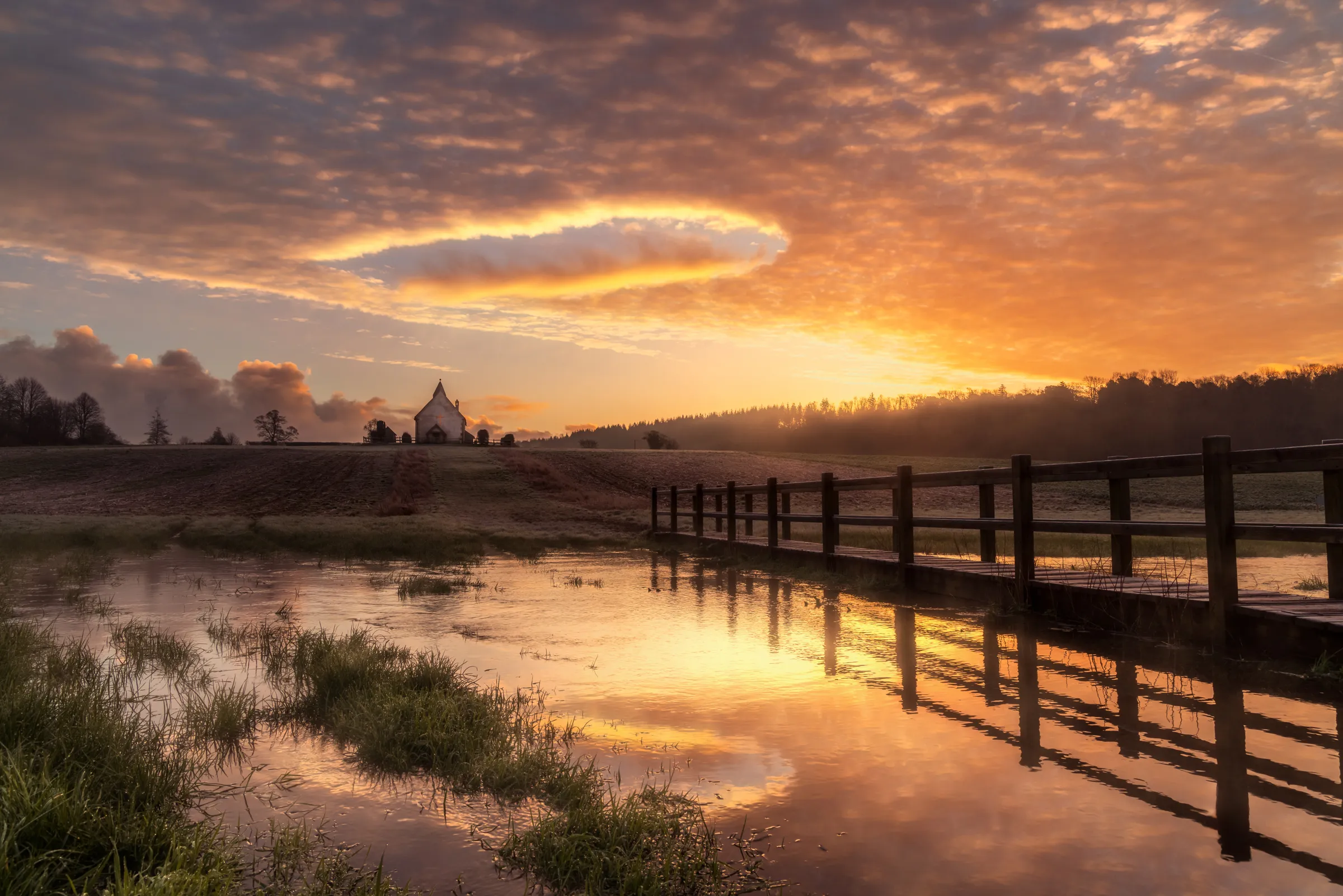 St Hubert's Chapel at Idsworth at sunrise with a circular cloud formation above, reflected in floodwater beside a wooden footbridge.