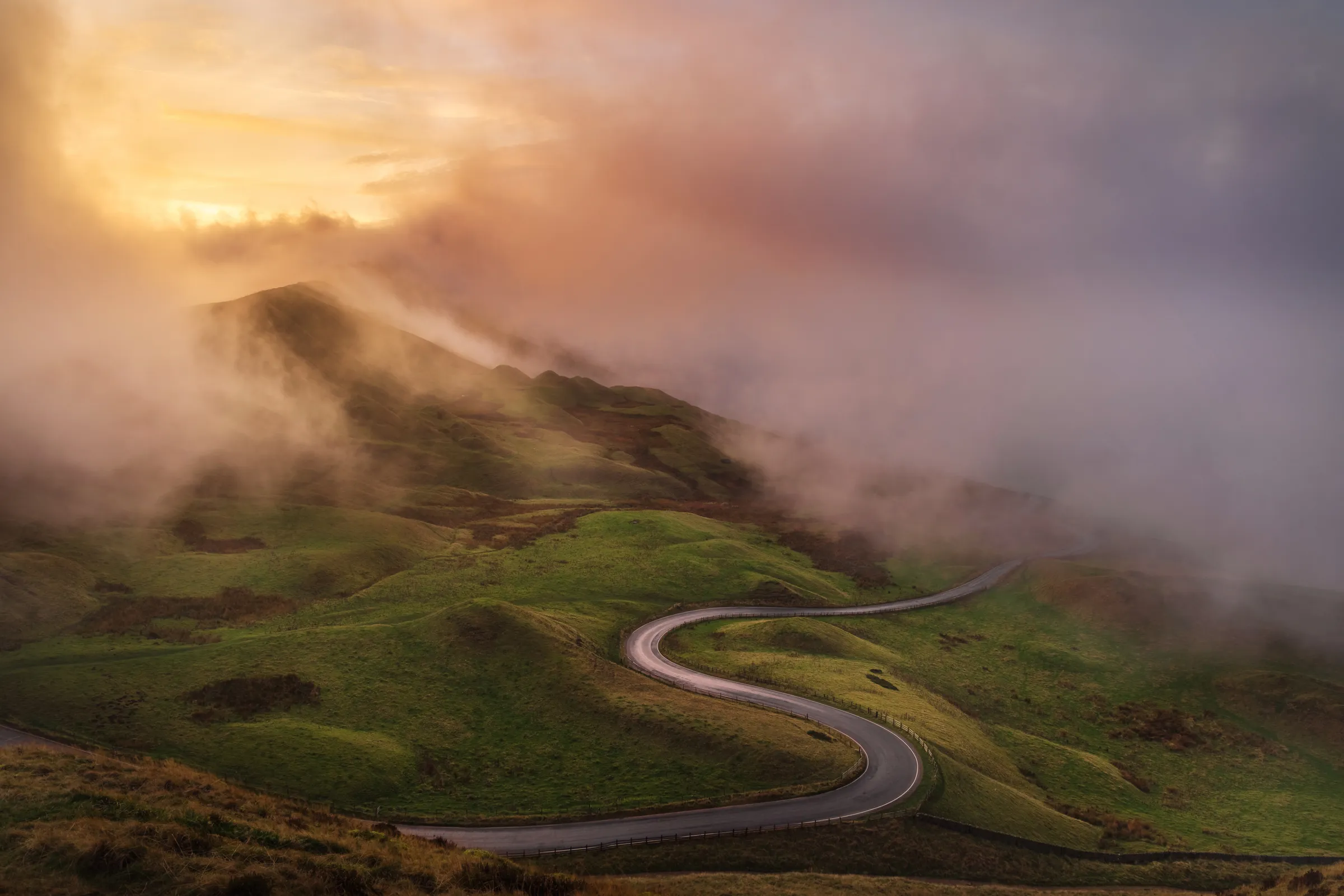 A winding road cuts through mist-covered hills in the Peak District at sunset, with low cloud drifting across the landscape and soft warm light breaking through.