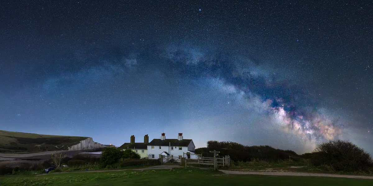 The Milky Way arching over the Coastguard Cottages at Cuckmere Haven, with the Seven Sisters cliffs visible beneath a star-filled sky.
