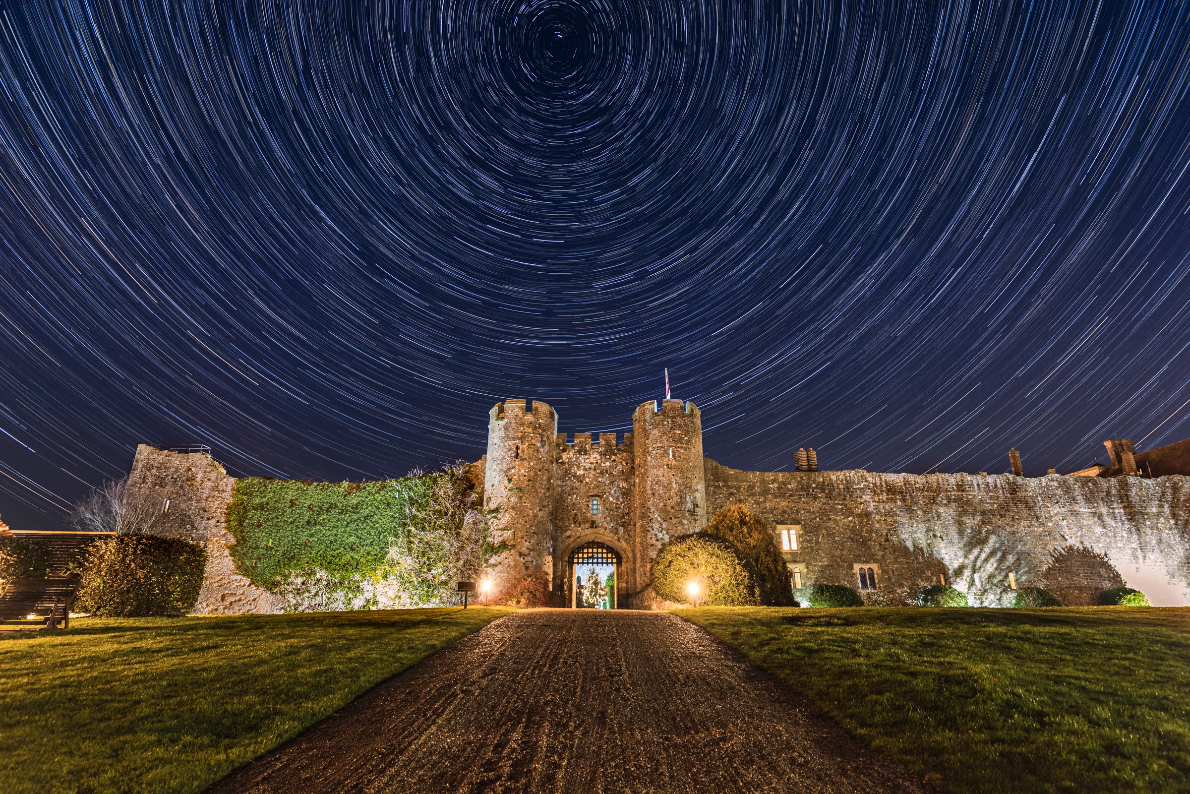 Amberley Castle gate beneath concentric star trails, with circular arcs radiating around the celestial pole above the historic walls.