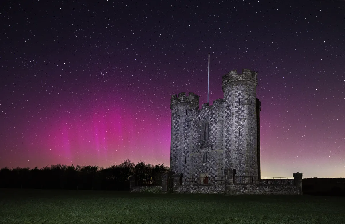 Hiorne Tower illuminated beneath a star-filled sky, with a crimson aurora glow spreading across the horizon behind the historic structure.