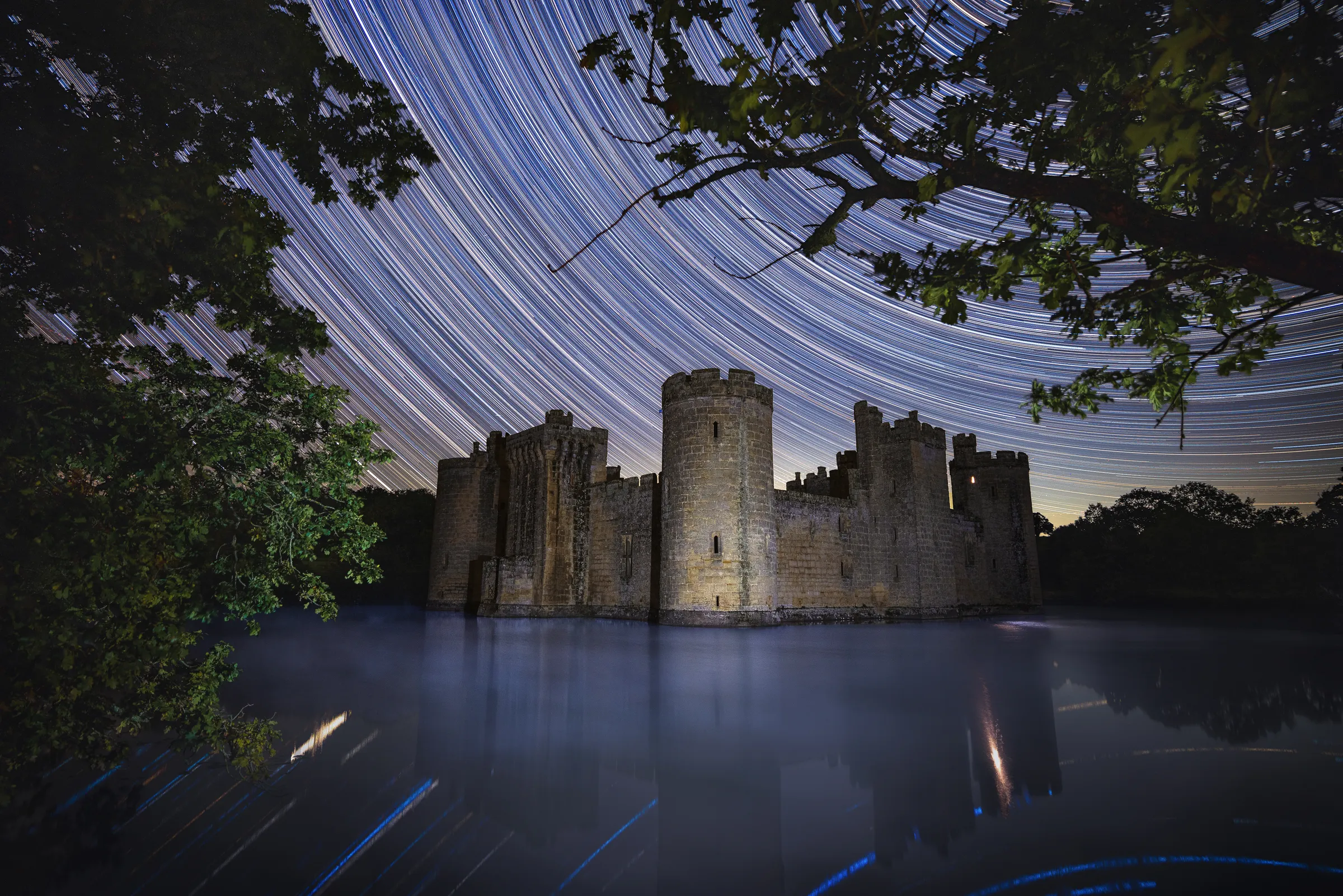 Bodiam Castle surrounded by water beneath sweeping star trails, with arcs stretching across the sky above the illuminated fortress.