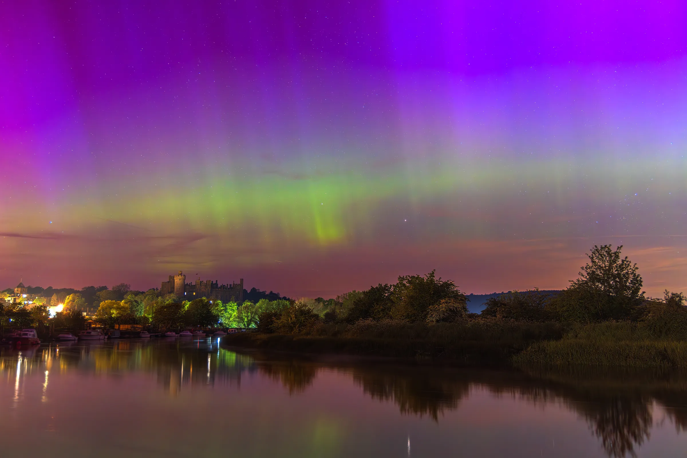 Arundel Castle beneath a vivid aurora, with green and purple light stretching across the sky and reflecting in the River Arun below.