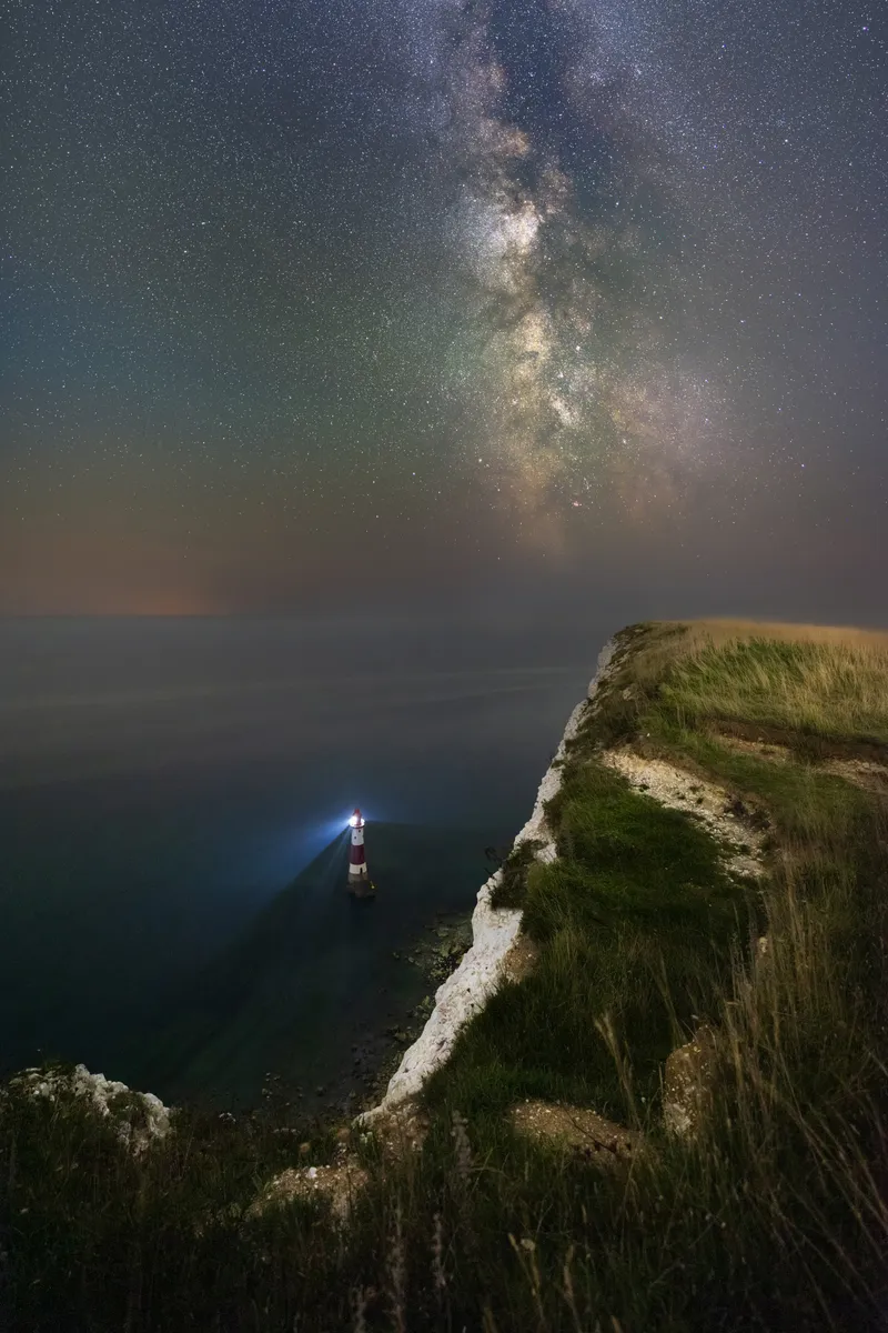 Beachy Head Lighthouse beneath the Milky Way, viewed from the cliffs above with the galaxy rising vertically into a star-filled sky.