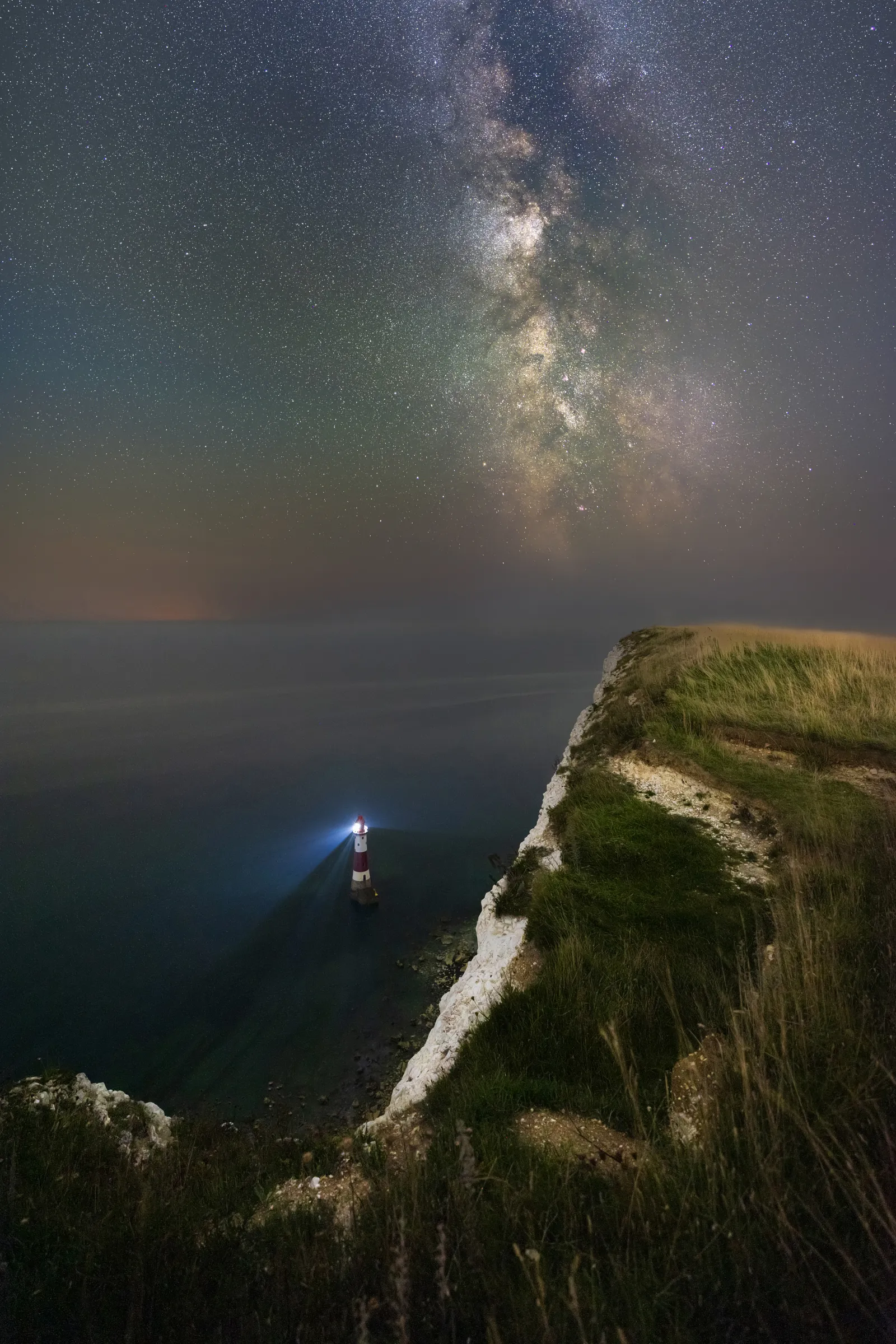 Beachy Head Lighthouse beneath the Milky Way, viewed from the cliffs above with the galaxy rising vertically into a star-filled sky.