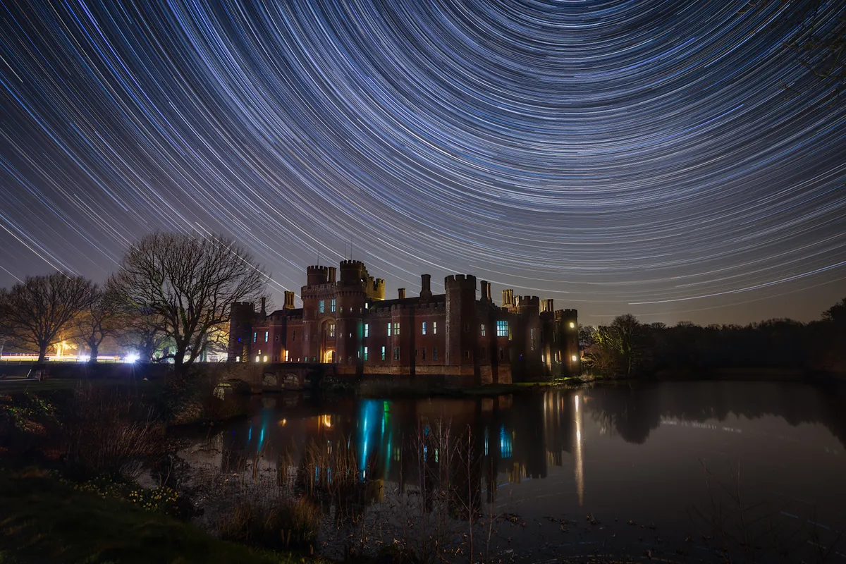 Herstmonceux Castle reflected in still water beneath sweeping star trails, with circular arcs filling the night sky above the illuminated structure.