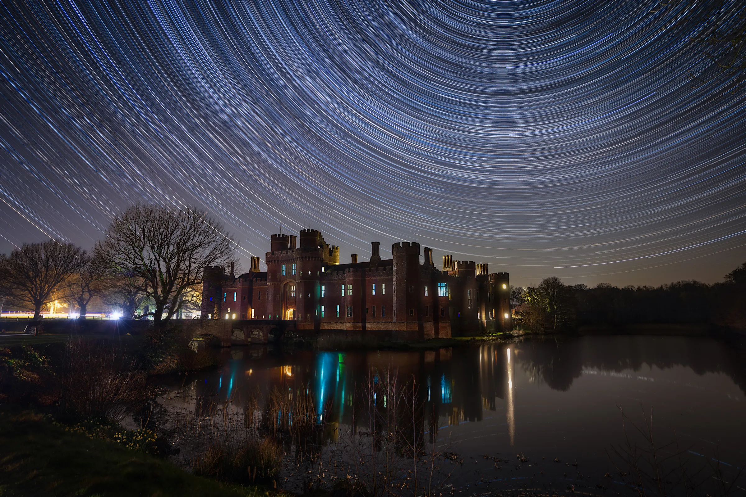 Herstmonceux Castle reflected in still water beneath sweeping star trails, with circular arcs filling the night sky above the illuminated structure.