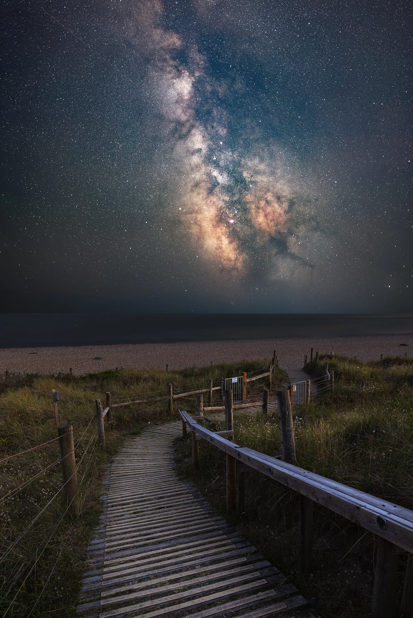 A wooden boardwalk leading through dunes toward the sea beneath the Milky Way, with a dense star field stretching across the night sky.