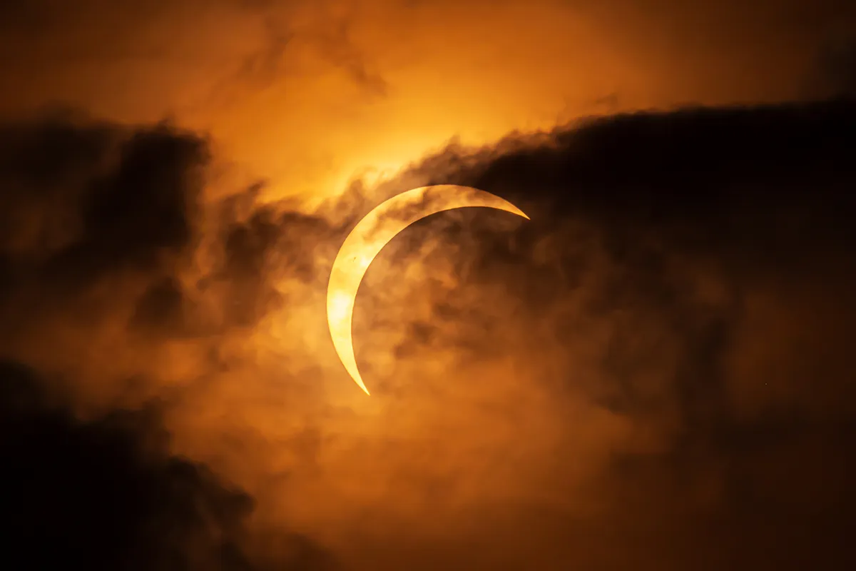 A crescent Sun during the August 2026 solar eclipse shining through dramatic layers of cloud above Talacre Beach in North Wales.
