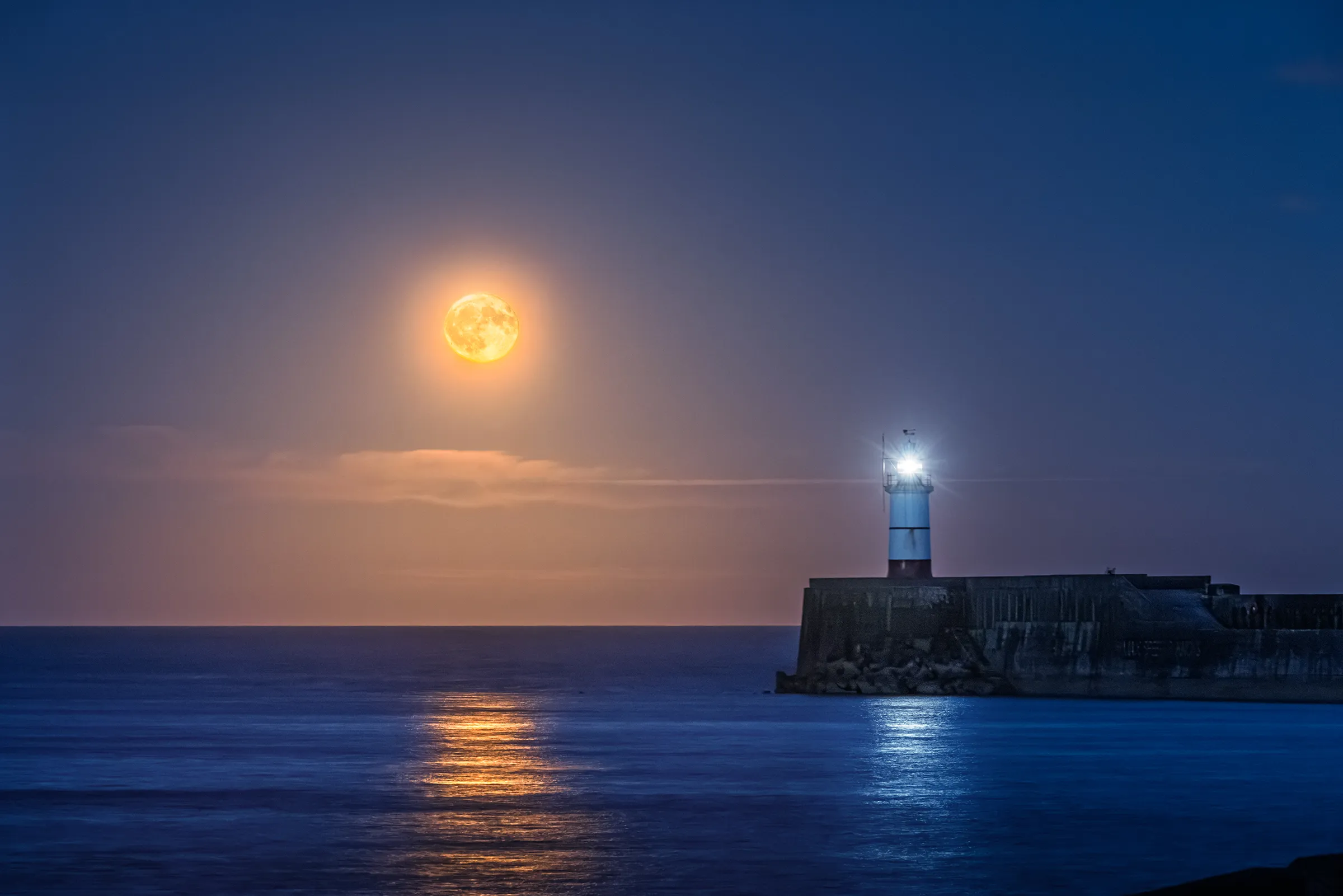 The Strawberry Moon rises above the sea beside Newhaven Lighthouse, with warm moonlight and cool lighthouse reflections across the water.