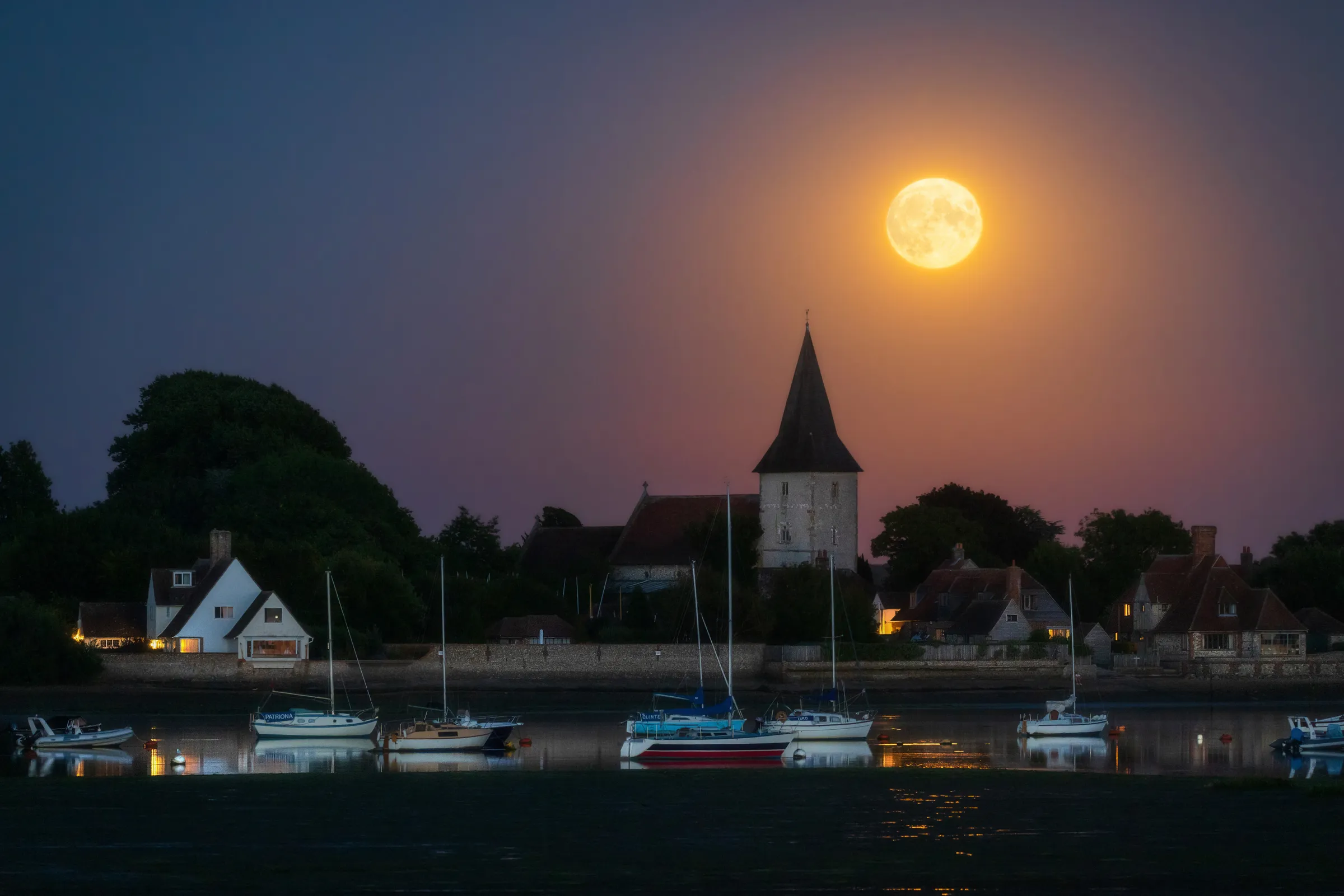 A full Sturgeon Moon rising above Holy Trinity Church in Bosham, with boats resting on calm water in the foreground.