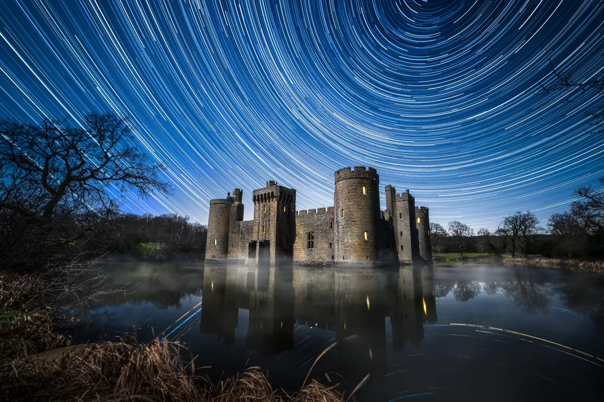 Bodiam Castle reflected in still water beneath sweeping star trails, with arcs curving around the sky above the illuminated fortress.