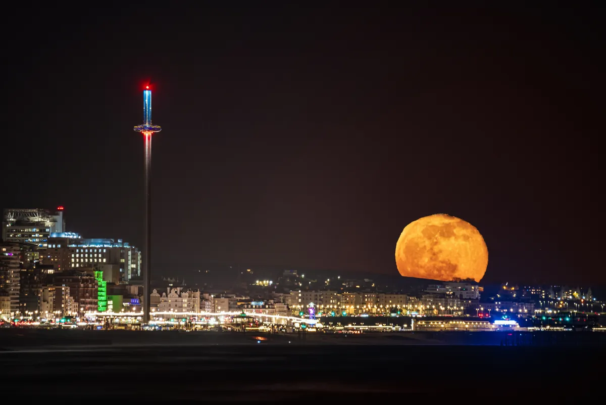A large orange full moon rising over Brighton seafront with the i360 tower and city lights visible along the coastline.
