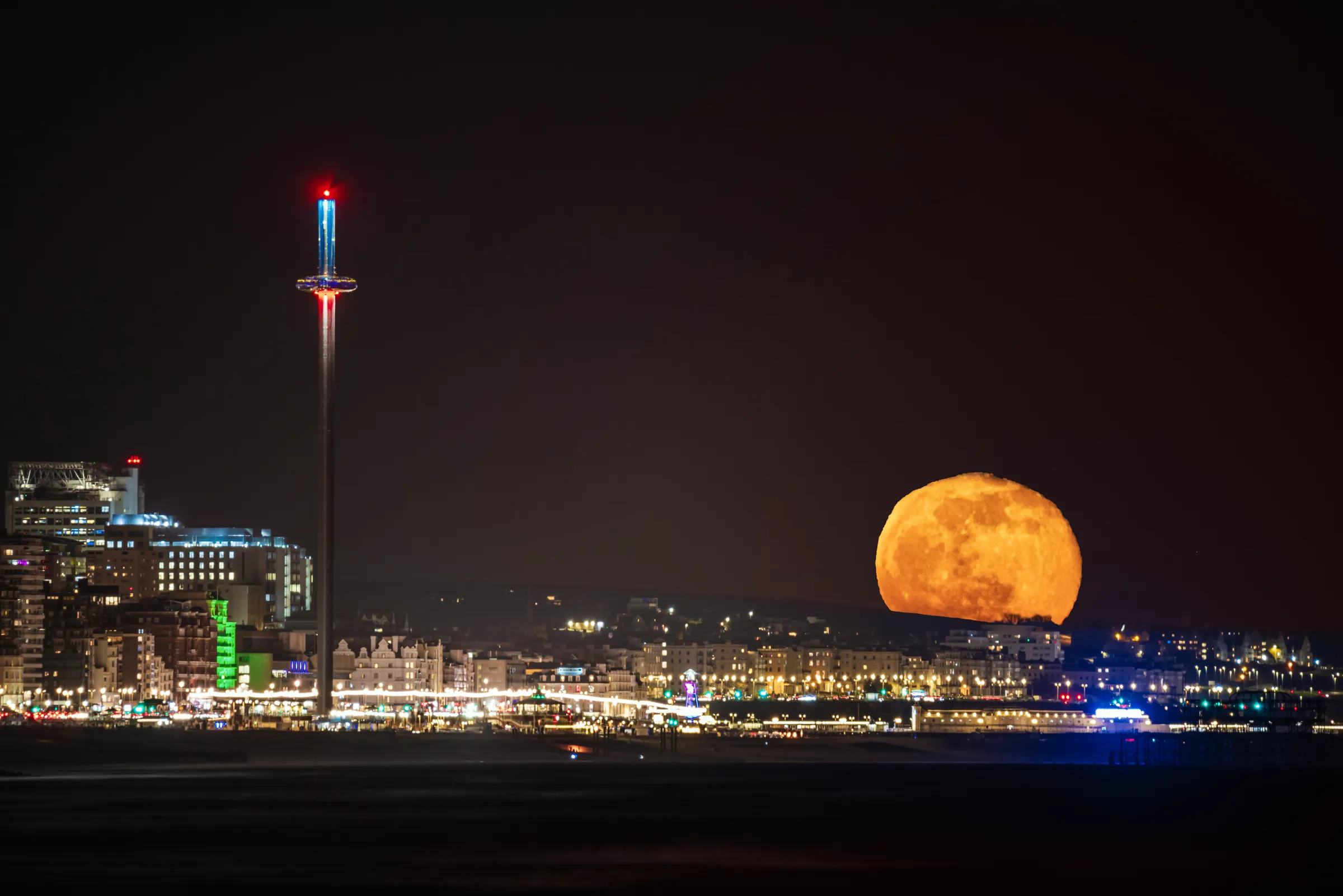 A large orange full moon rising over Brighton seafront with the i360 tower and city lights visible along the coastline.