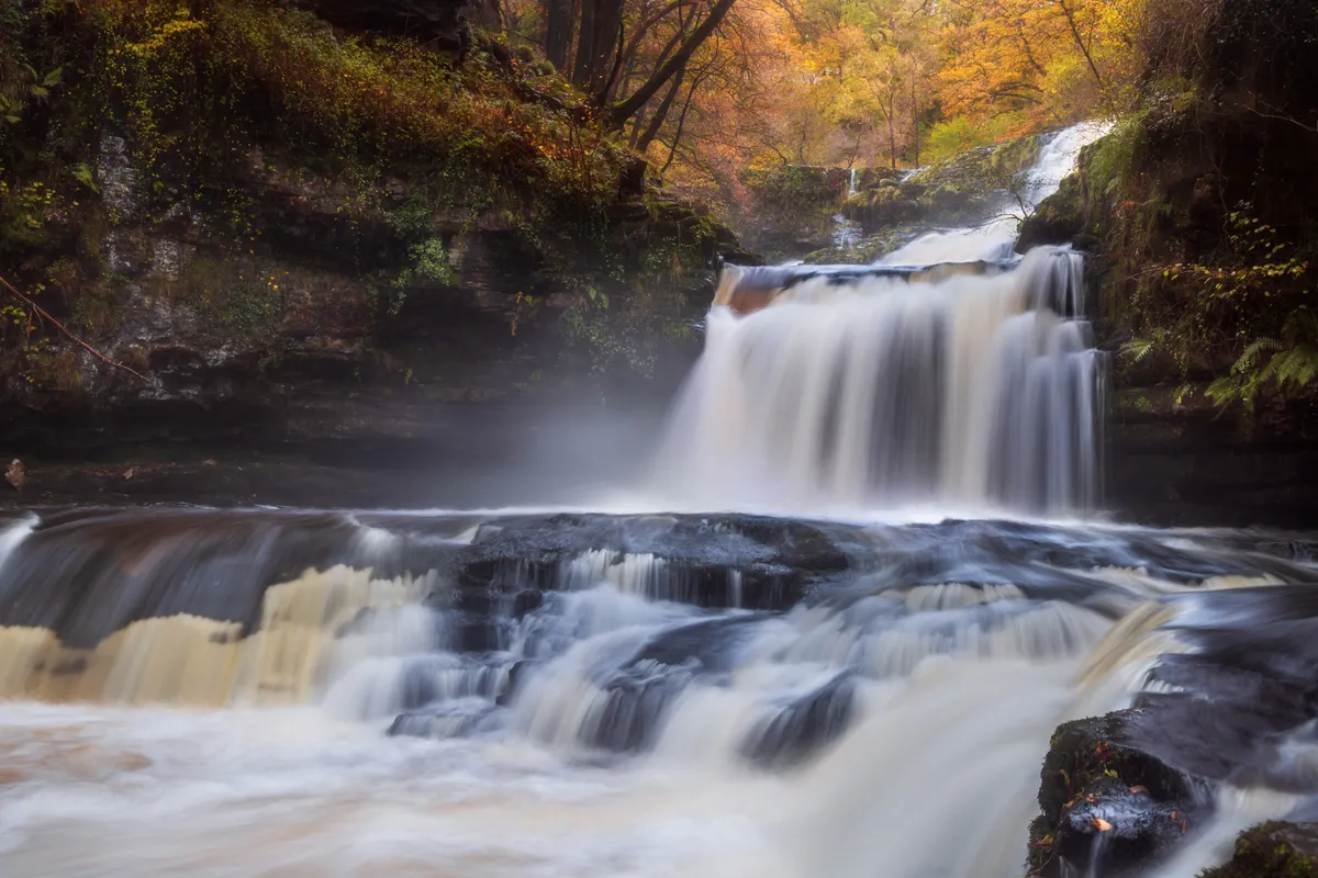 Isaf Clun Gwyn waterfall cascading over rock in the Brecon Beacons, surrounded by autumn foliage and soft mist.