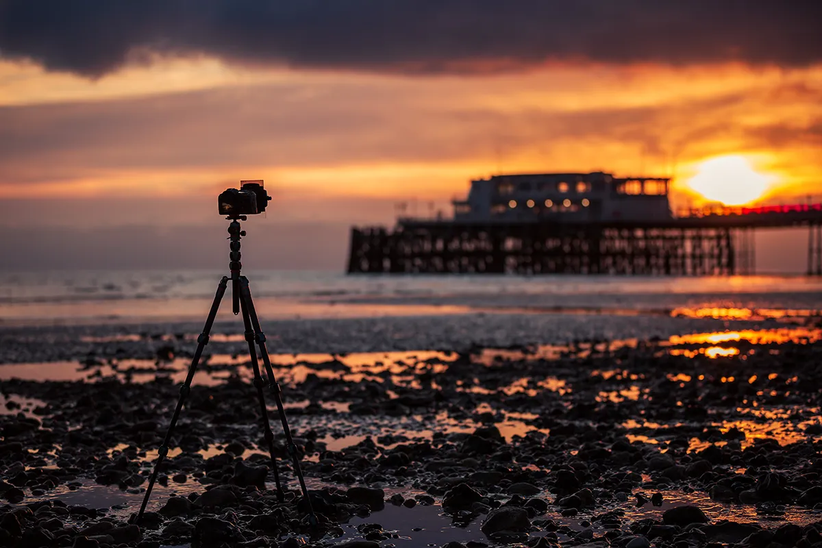 A camera on a tripod silhouetted against a vivid sunset sky, with a pier visible in the background