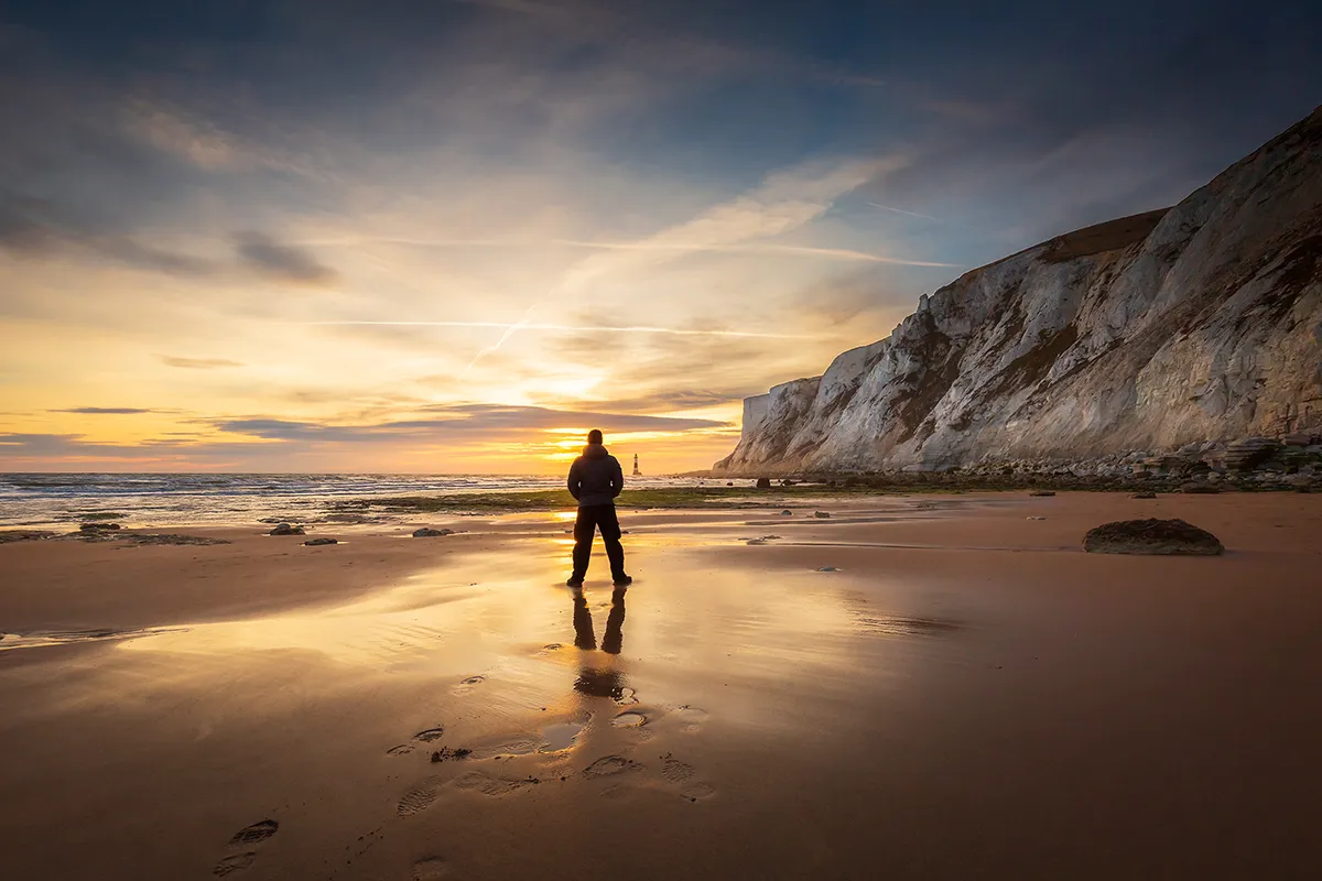 Stewart Reading-Brown standing on a beach at sunset, white chalk cliffs and a distant lighthouse behind him